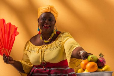 Gülümseyen orta yaşlı Afrikalı Amerikalı kadın meyvelere dokunuyor ve turuncu yelpaze tutuyor.
