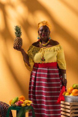 Pozitif orta yaşlı Afro-Amerikalı kadın elinde ananas ve vantilatörle meyve satıyor.