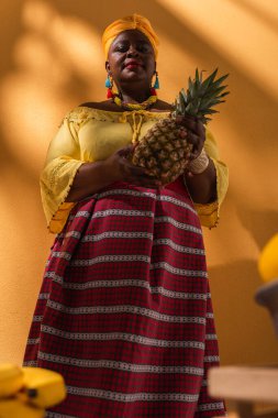 Düşük açı görüşlü orta yaşlı Afro-Amerikalı kadın elinde ananas ve portakal satan kadın.