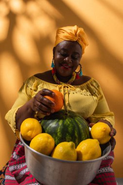 smiling middle aged african american woman holding metal bowl with watermelon and fruits on orange clipart