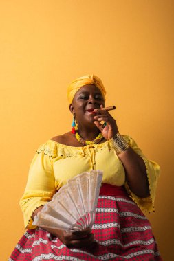 middle aged african american woman in bright clothes sitting with fan and smoking cigar on yellow clipart