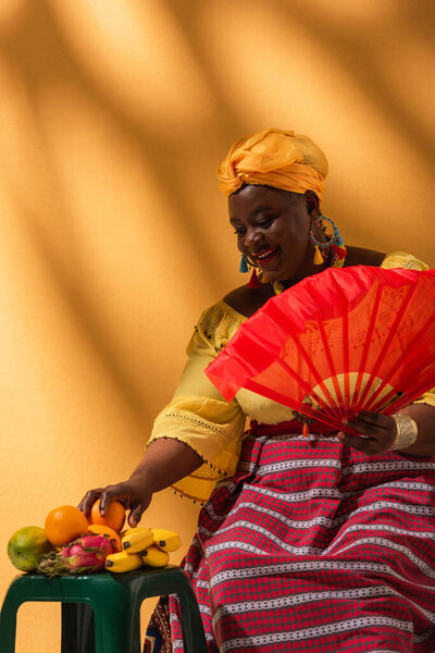 cheerful middle aged african american woman touching fruits and holding fan on yellow