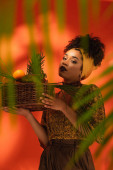 mladá africká americká žena drží koš s exotickým ovocem za rozmazané palmové listy na oranžové