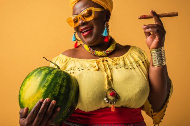 Orta yaşlı, parlak elbiseli, güneş gözlüklü, sarıda karpuz ve puro tutan Afrikalı Amerikalı kadın.