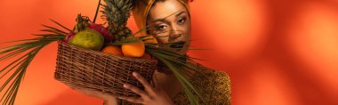 young african american woman holding basket with exotic fruits near face on orange, banner clipart