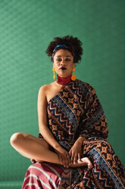 Afro-Amerikan kadın süs battaniyesine sarılmış yeşil desenli arka plandaki kameraya bakıyor.