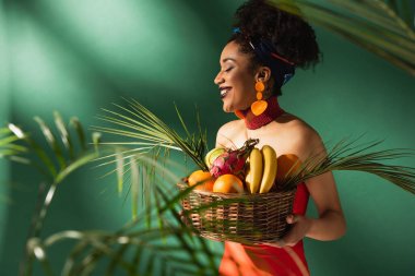 smiling young african american woman in red swimsuit holding basket with exotic fruits on green clipart