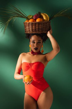 Kırmızı mayo giymiş, egzotik meyvelerle dolu yeşil sepetli genç Afro-Amerikan kadın.