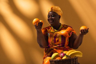 Pozitif orta yaşlı Afrikalı Amerikalı satış elemanı elinde portakal tutuyor ve arka planda gölgeli meyveler satıyor.
