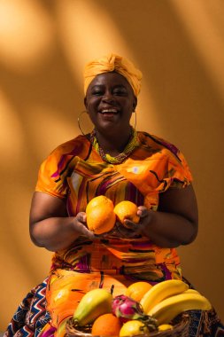 Gülümseyen orta yaşlı Afrikalı Amerikalı satıcı elinde meyvelerle turuncu üzerinde oturuyor.