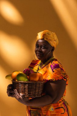smiling middle aged african american woman holding basket with exotic fruits on orange clipart