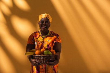 smiling middle aged african american woman in bright dress and turban holding basket with exotic fruits on orange clipart