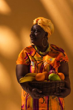 smiling middle aged african american woman holding basket with exotic fruits on orange clipart