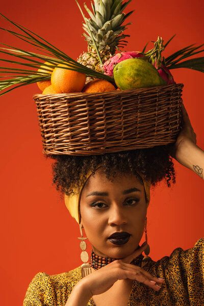 портрет серьезной молодой африканской женщины с корзиной экзотических фруктов на голове на красном