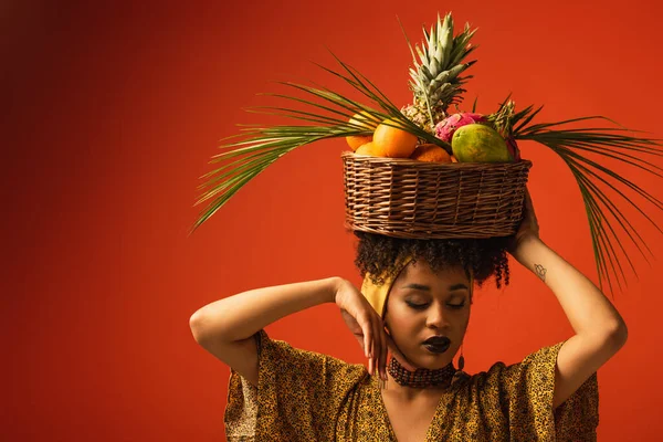 young african american woman with hand near face holding basket with exotic fruits on head on red