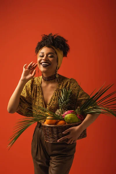 positive young african american woman with hand near face holding basket with exotic fruits on red