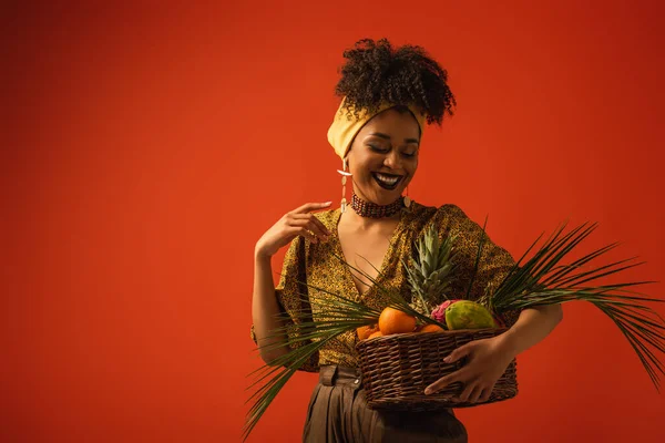 smiling young african american woman with hand near shoulder holding basket with exotic fruits on red