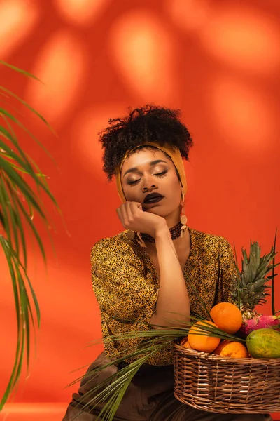 young african american woman with closed eyes and hand near face sitting near basket with exotic fruits on orange