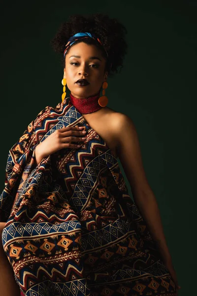 african american woman in colorful headscarf, earrings and necklace sitting wrapped in ornament blanket on dark green