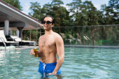 šťastný muž bez košile ve slunečních brýlích relaxace v bazénu s lahví piva