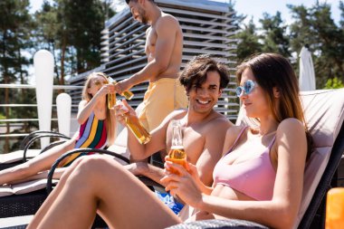 Mayolu arkadaşlar konuşuyor ve tatil köyünde bira şişeleri tutuyorlar. 