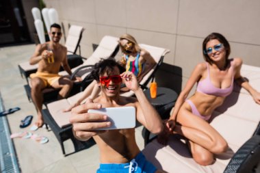 Akıllı telefon elinde. Arkadaşlarının yanında mayo ve sandalyelerle selfie çekiyor. 