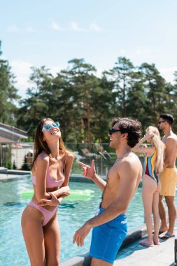 Yüzme şortlu neşeli adam dışarıdaki havuzun yanındaki mayo giymiş arkadaşını işaret ediyor. 