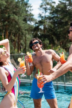 Happy man with cocktail standing near multiethnic friends and swimming pool during weekend  clipart