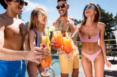 Cocktails in hands of cheerful interracial friends near blurred swimming pool  clipart