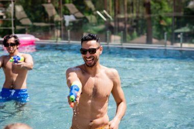 cheerful muslim man playing with water gun near blurred friend in pool clipart