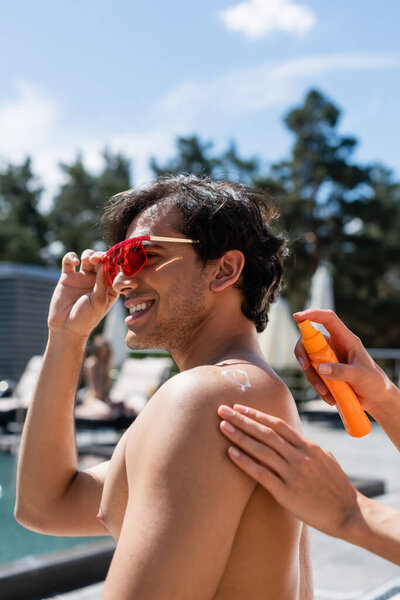 Woman applying sunscreen on back of smiling friend in sunglasses