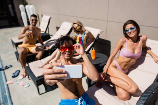 Smartphone in hand of man taking selfie near friends in swimwear on deck chairs 