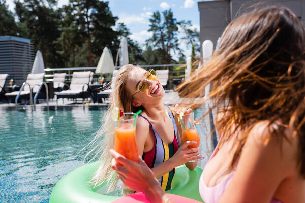 счастливая женщина с коктейльным бокалом смеется рядом с размытым другом в бассейне