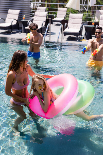 blurred interracial friends playing with water pistols near laughing women in swimming pool