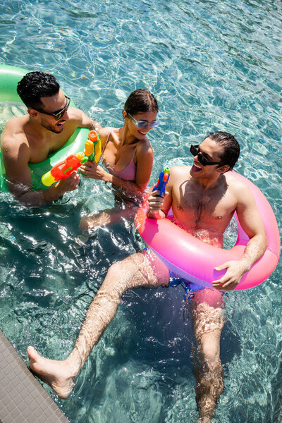 cheerful multicultural friends relaxing in pool with water guns and swim rings