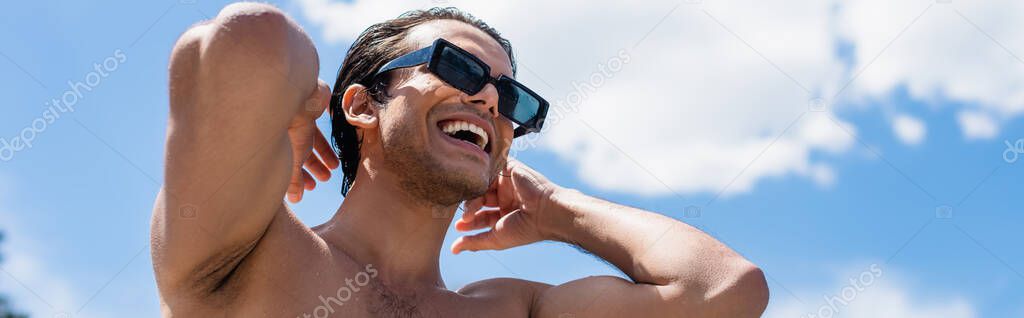 low angle view of young shirtless man in sunglasses smiling against blue sky, banner