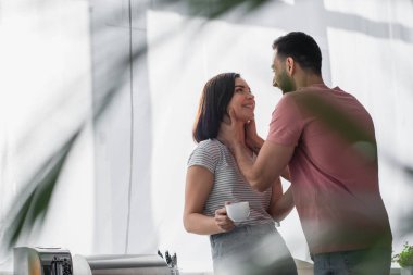 Gülümseyen genç adam modern mutfakta beyaz kahve fincanıyla kız arkadaşının boynuna dokunuyor.
