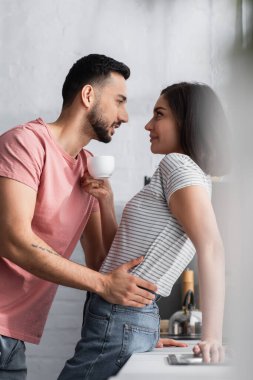 Genç aşıklar modern mutfakta kahve fincanıyla sarılıyorlar.