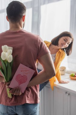 Genç bir adamın elinde çiçek buketi ve bulanık kız arkadaşının yanında tebrik kartı tutuşu.