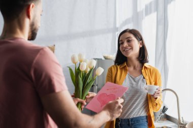 Genç adam mutfaktaki gülümseyen kız arkadaşına bir buket çiçek ve tebrik kartı sunuyor.