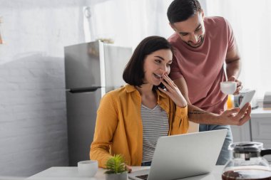 Gülümseyen genç adam evde dizüstü bilgisayarla kız arkadaşına cep telefonunu gösteriyor.