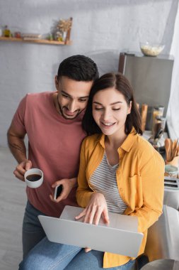 Gülümseyen genç adam cep telefonu ve kahve bardağıyla kız arkadaşının yanında dizüstü bilgisayarla evde oturuyor.