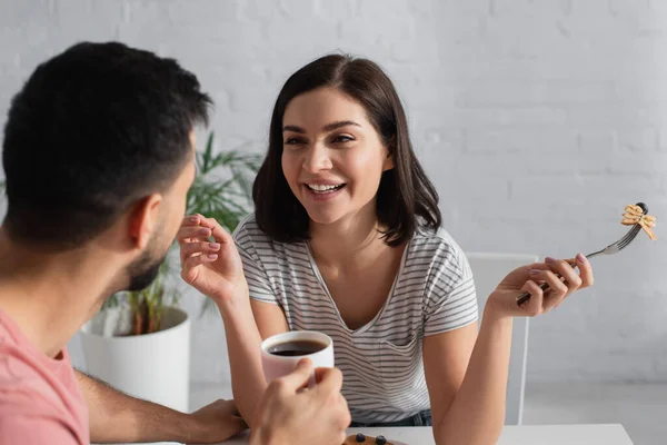 Leende Ung Kvinna Äter Pannkakor Nära Suddig Pojkvän Med Kaffekopp — Stockfoto