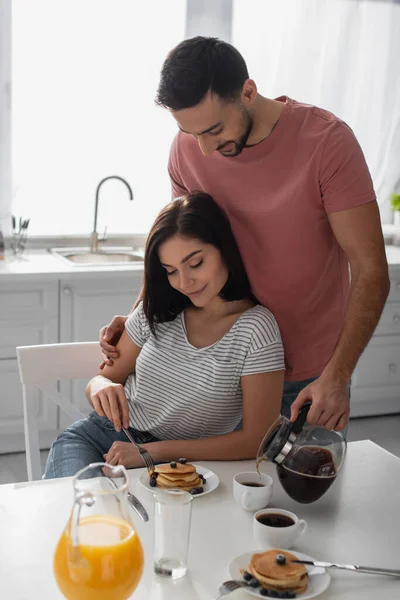 Smiling Young Man Hugging Girlfriend Eating Pancakes Pouring Coffee Pot — Stock Photo, Image