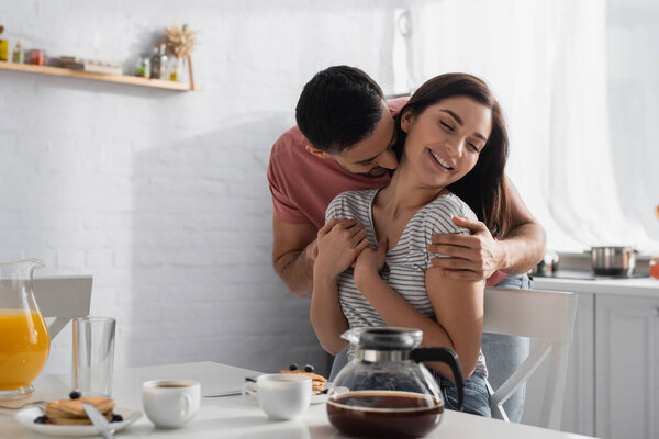 happy young couple hugging and kissing near table with breakfast, coffee and orange juice in kitchen