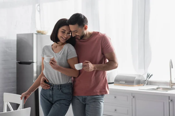 Pasangan Muda Tersenyum Memeluk Dengan Cangkir Kopi Dapur — Stok Foto