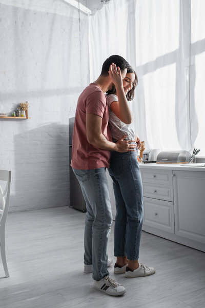 позитивная молодая пара в повседневной одежде нежно обнимается и танцует на современной кухне