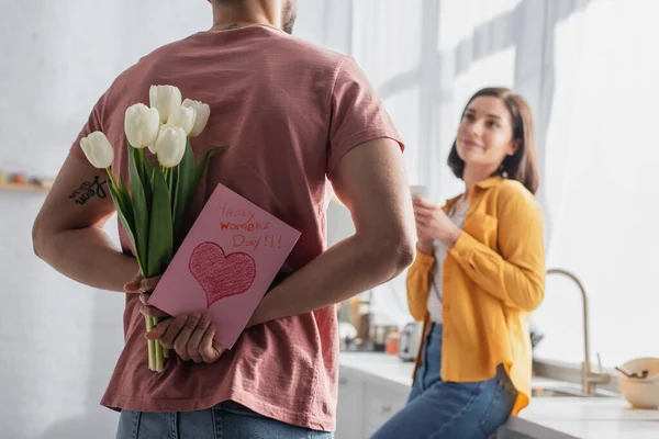 在厨房里 一个年轻人拿着一束鲜花和一张贺卡站在模糊的女朋友旁边 他的倒影映入眼帘 — 图库照片
