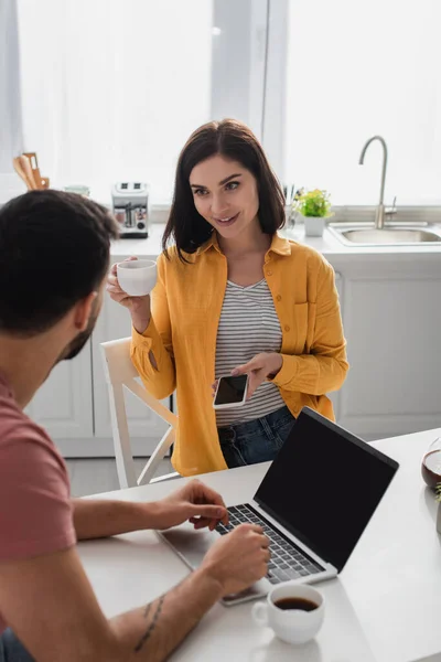 Sonriente Joven Bebiendo Café Sosteniendo Teléfono Celular Cerca Novio Trabajando — Foto de Stock