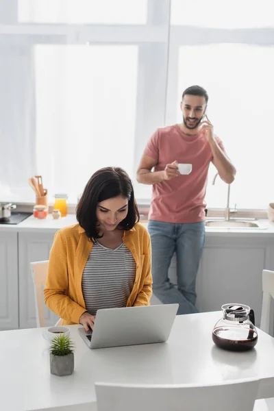 Smiling Young Woman Working Laptop Blurred Boyfriend Speaking Cellphone Kitchen — Stock Photo, Image